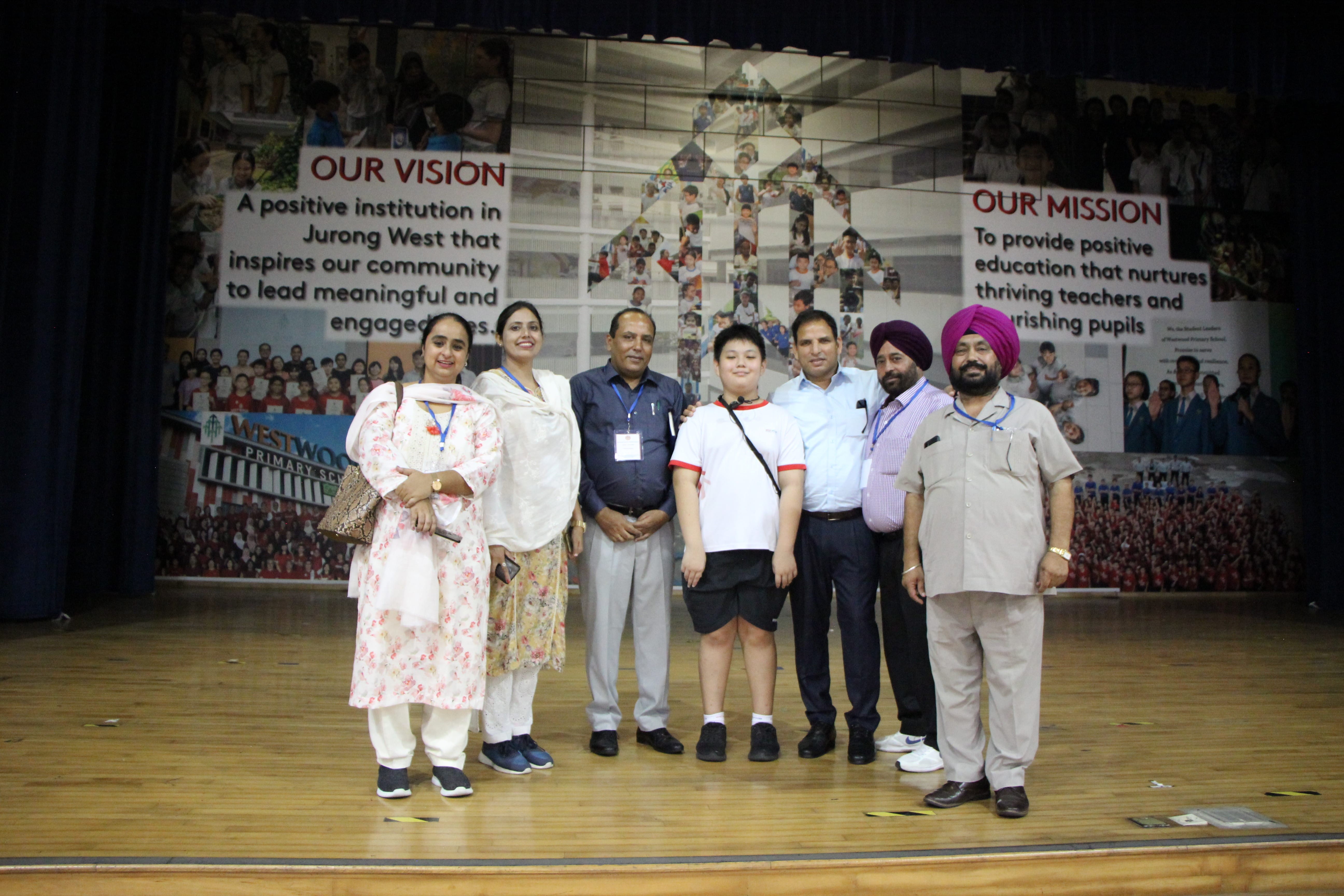 Six adults and one child pose on a stage in front of a Westwood Primary School vision and mission banner.